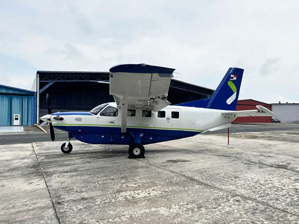 A blue and white Quest Kodiak 100 aircraft with a green tail logo parked on a concrete apron in front of a hangar.