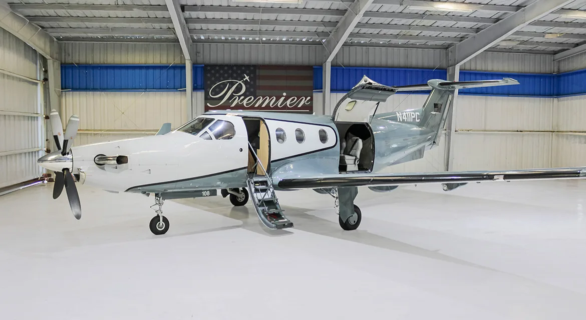 A white and grey Pilatus PC-12 turboprop aircraft, tail number N411PC, with its main cabin door and cargo door open, parked in a hangar.