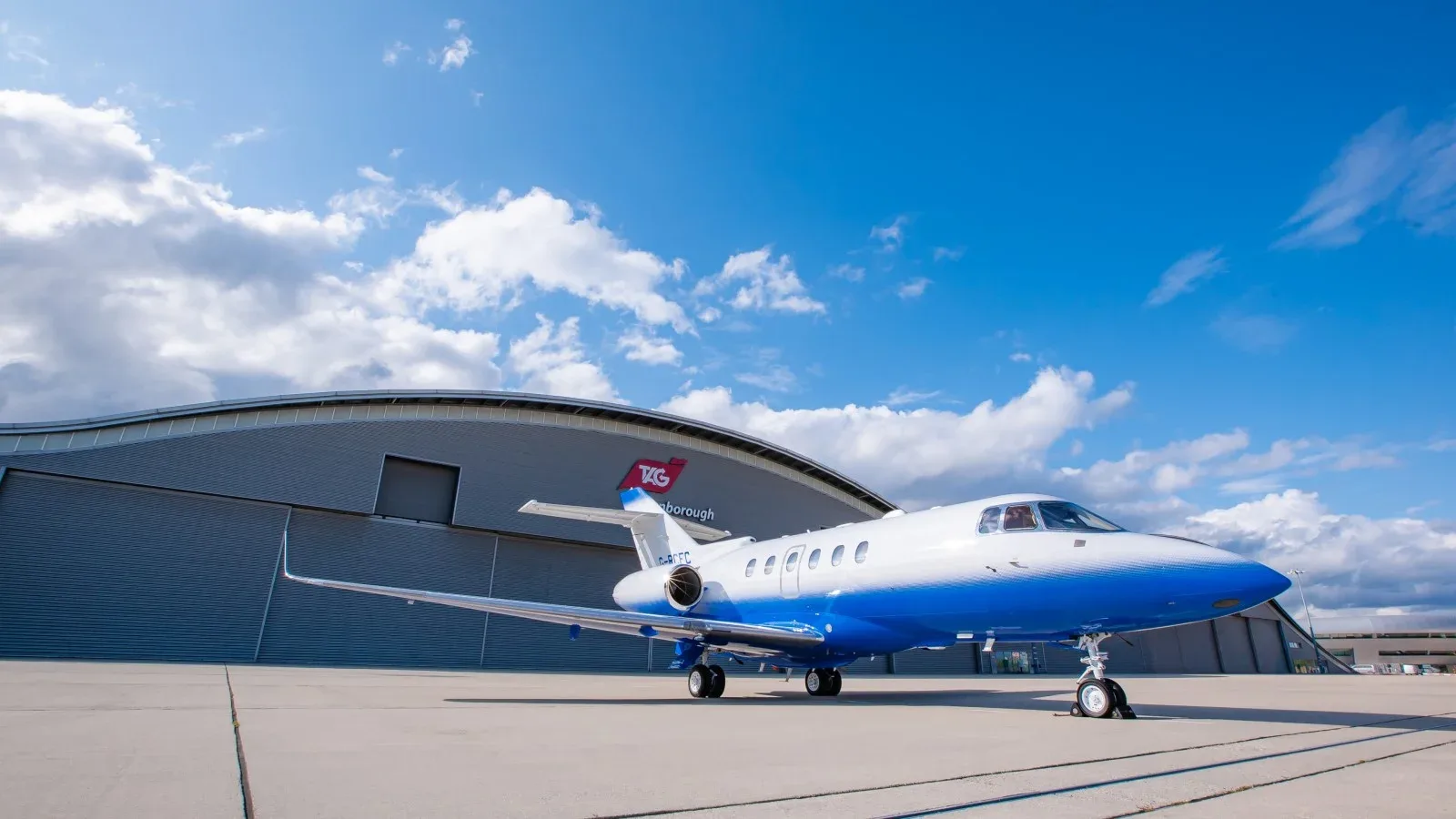 A blue and white jet sits on the tarmac in front of a hangar at Farnborough Airport.