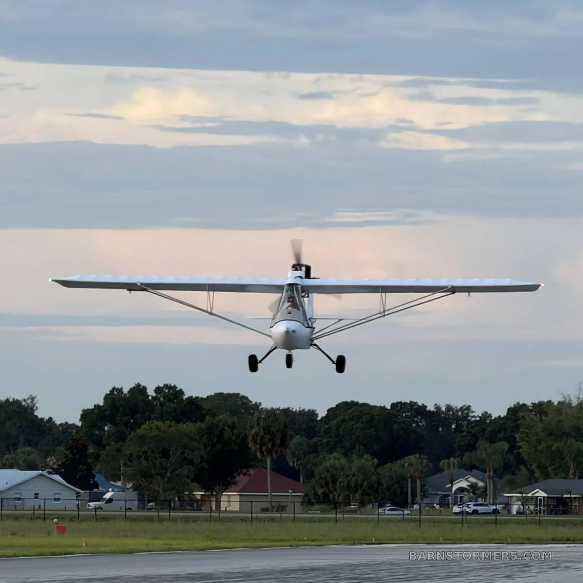 A white experimental ultralight aircraft with a high wing is seen from the front, just above a runway, with houses and trees in the background under a cloudy sky.