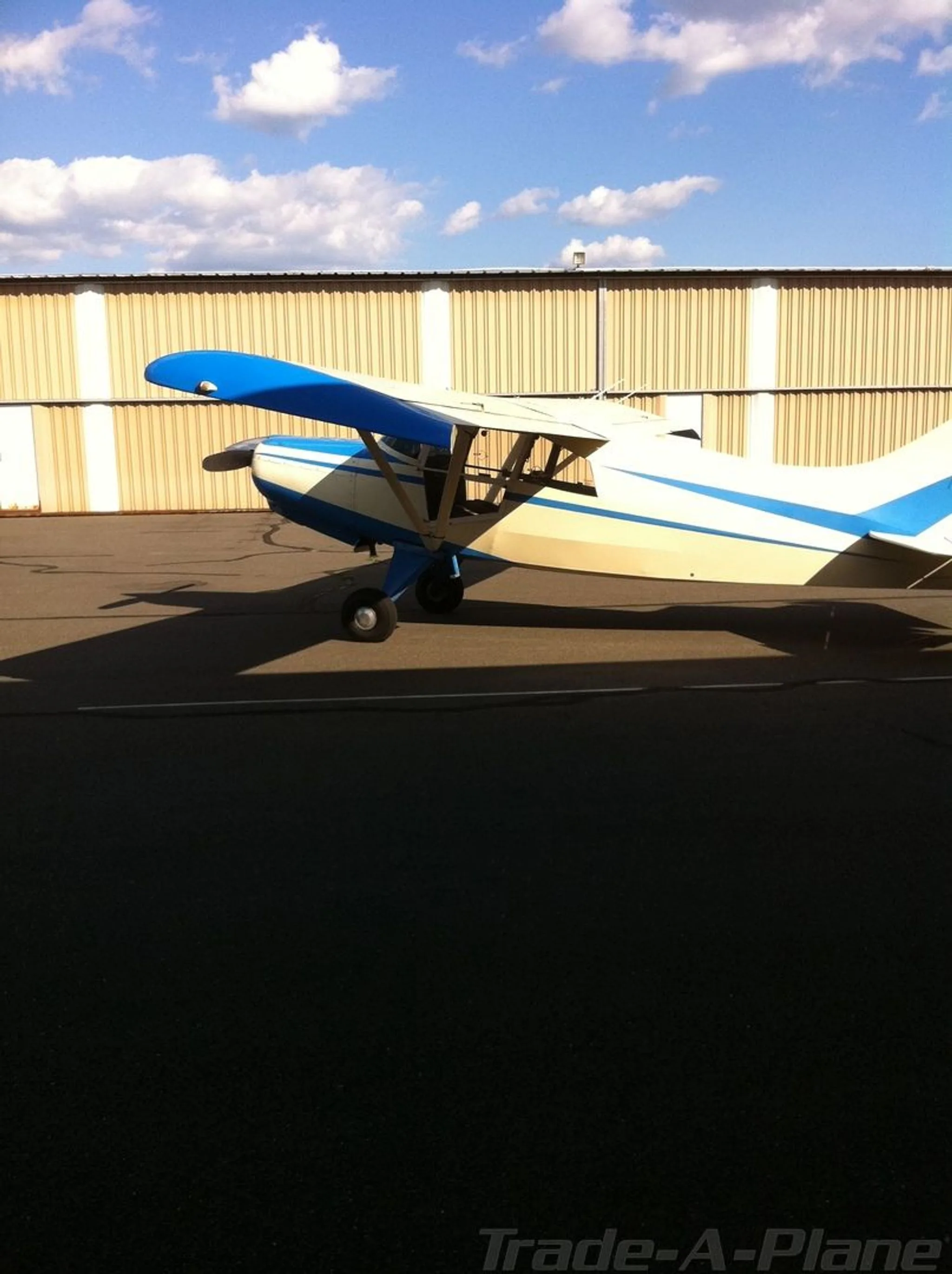 A white and blue high-wing taildragger aircraft is parked on an airfield, seen from the side, with a hangar in the background.