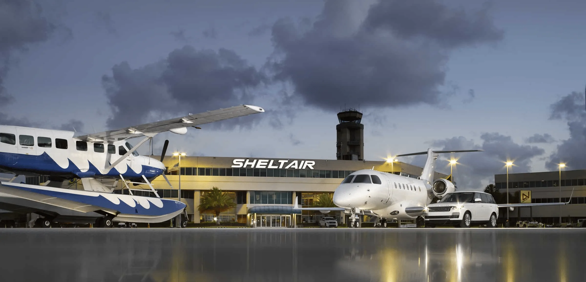 A white seaplane and a white business jet are parked on a wet tarmac in front of a SHELTAIR building and control tower at dusk.