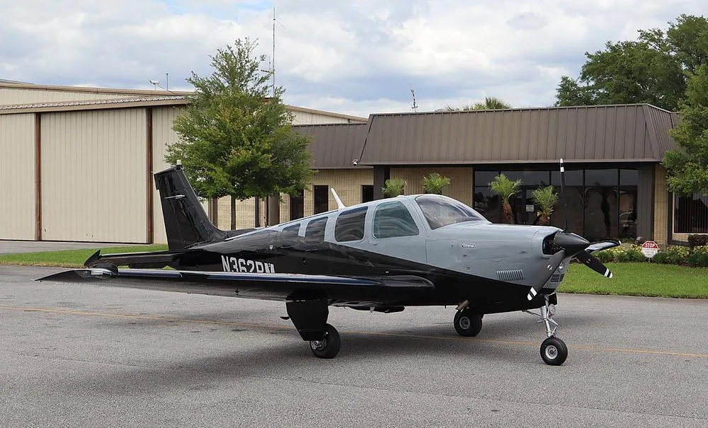 A black and grey single-engine propeller aircraft, registration N362RM, is parked on an asphalt ramp with hangars and trees in the background.