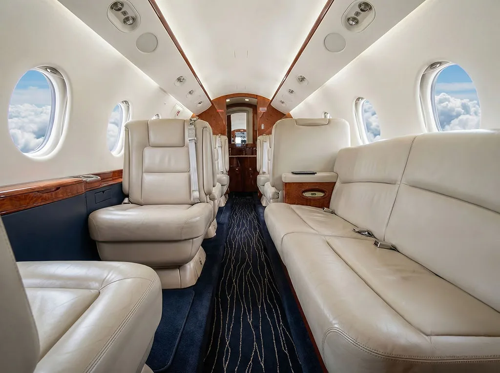 The private jet cabin features white leather seating, a sofa, and wooden accents, looking towards the rear.