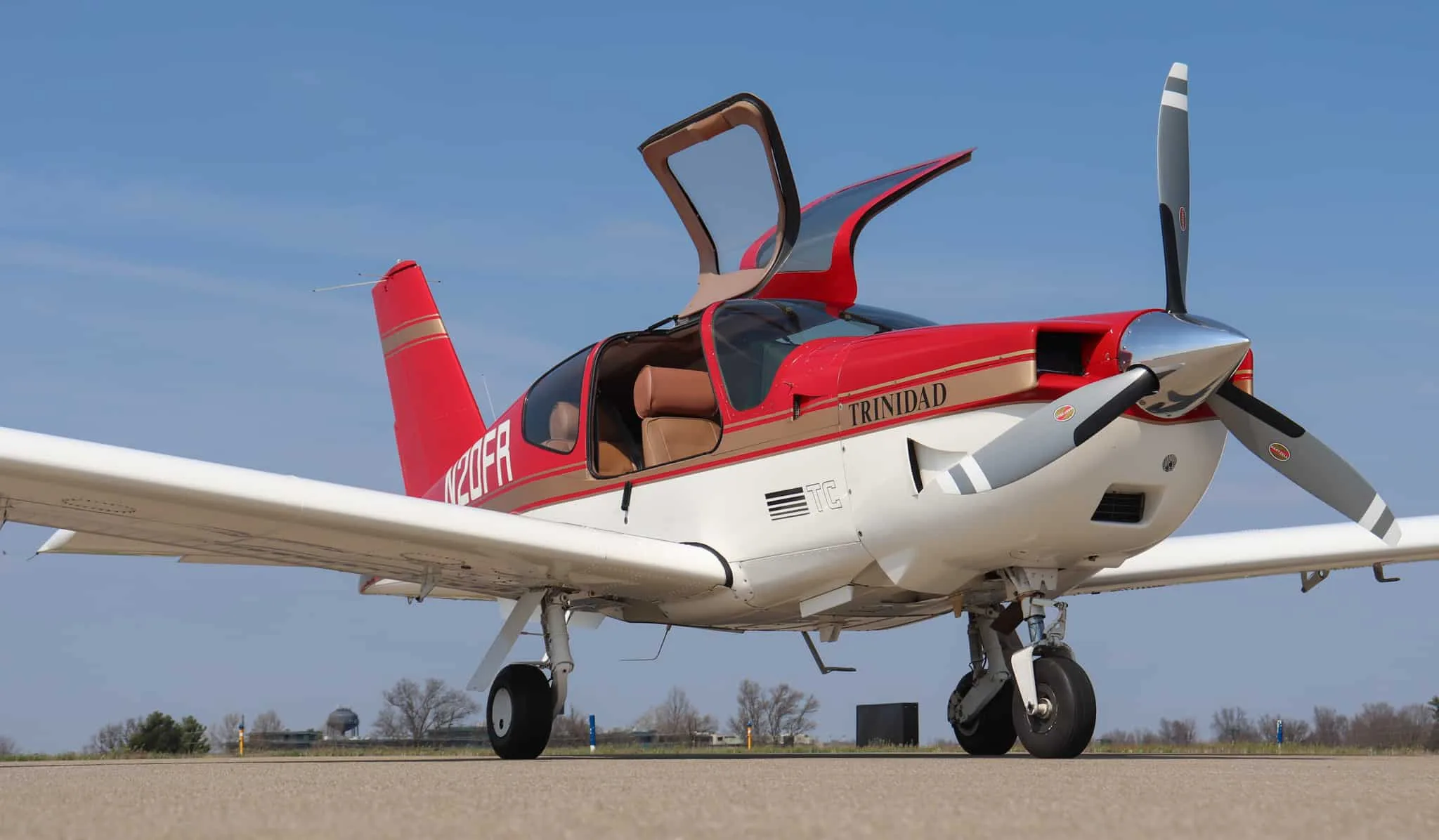A red and white Socata Trinidad aircraft with gold stripes and open doors is parked on an airport tarmac.