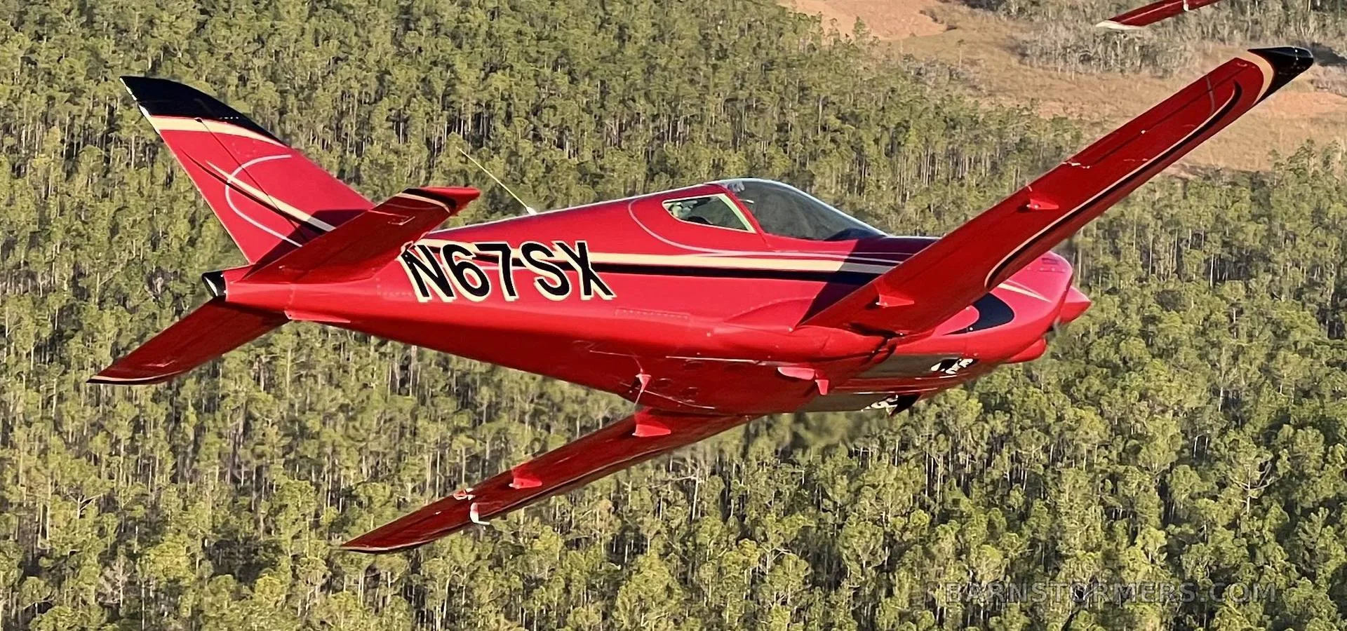 A red single-engine aircraft with registration N67SX is seen from behind and below, flying over a dense green forest.