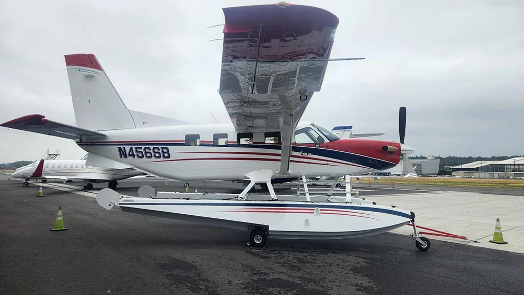 A side view of a red, white, and blue Quest Kodiak 100 on amphibious floats parked on an asphalt apron.