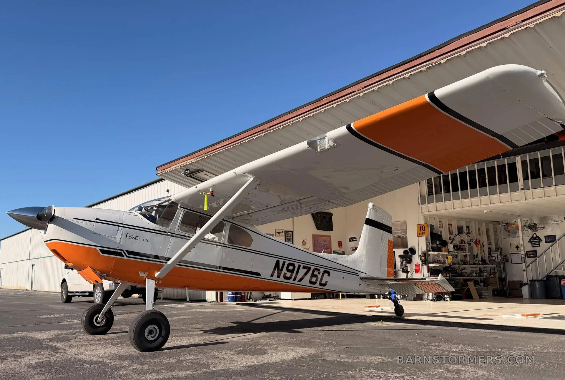 A white and orange Cessna 180 aircraft, registration N9176C, parked on an asphalt ramp next to a hangar under a clear blue sky.