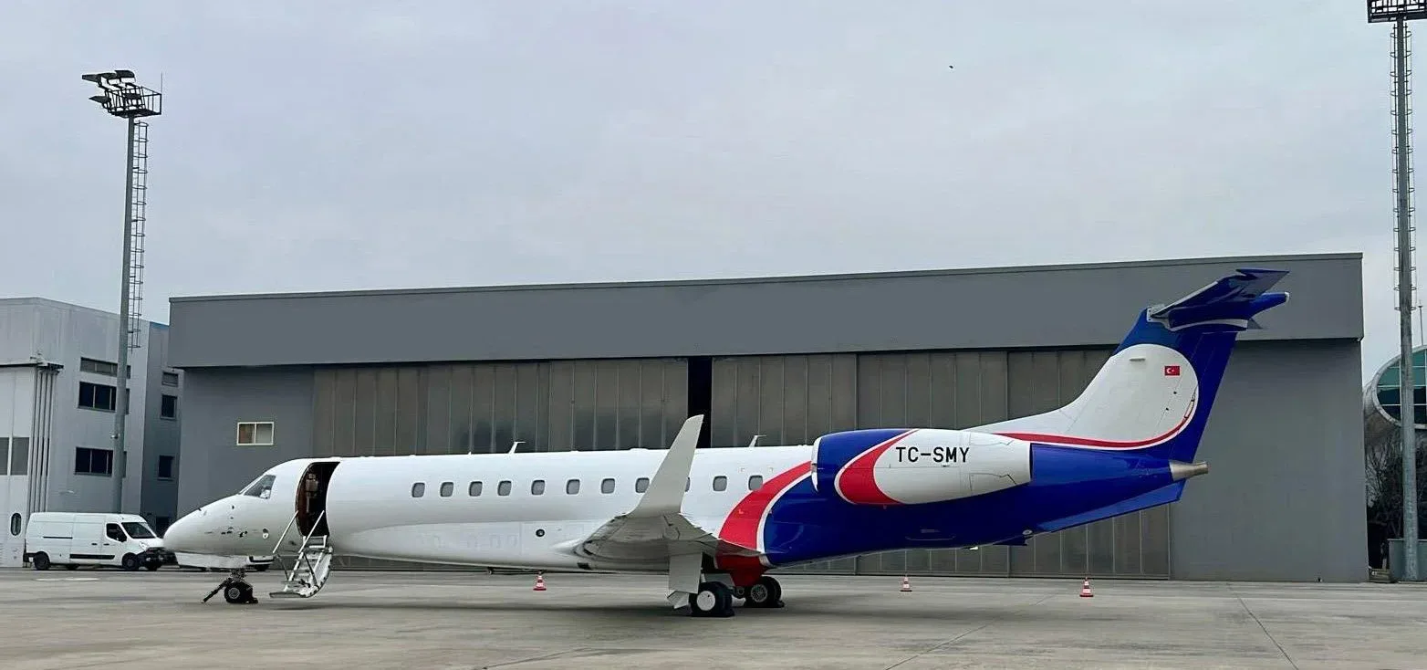 A white and blue Embraer Legacy 600 business jet with registration TC-SMY is parked on a tarmac next to a hangar.
