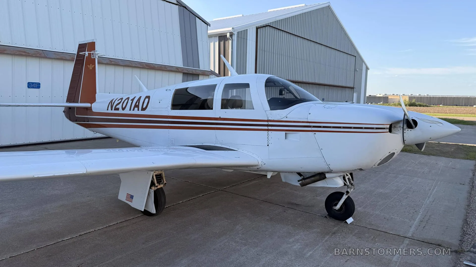 A white single-engine aircraft with brown stripes and tail, registration N201AD, parked on a tarmac in front of hangars.