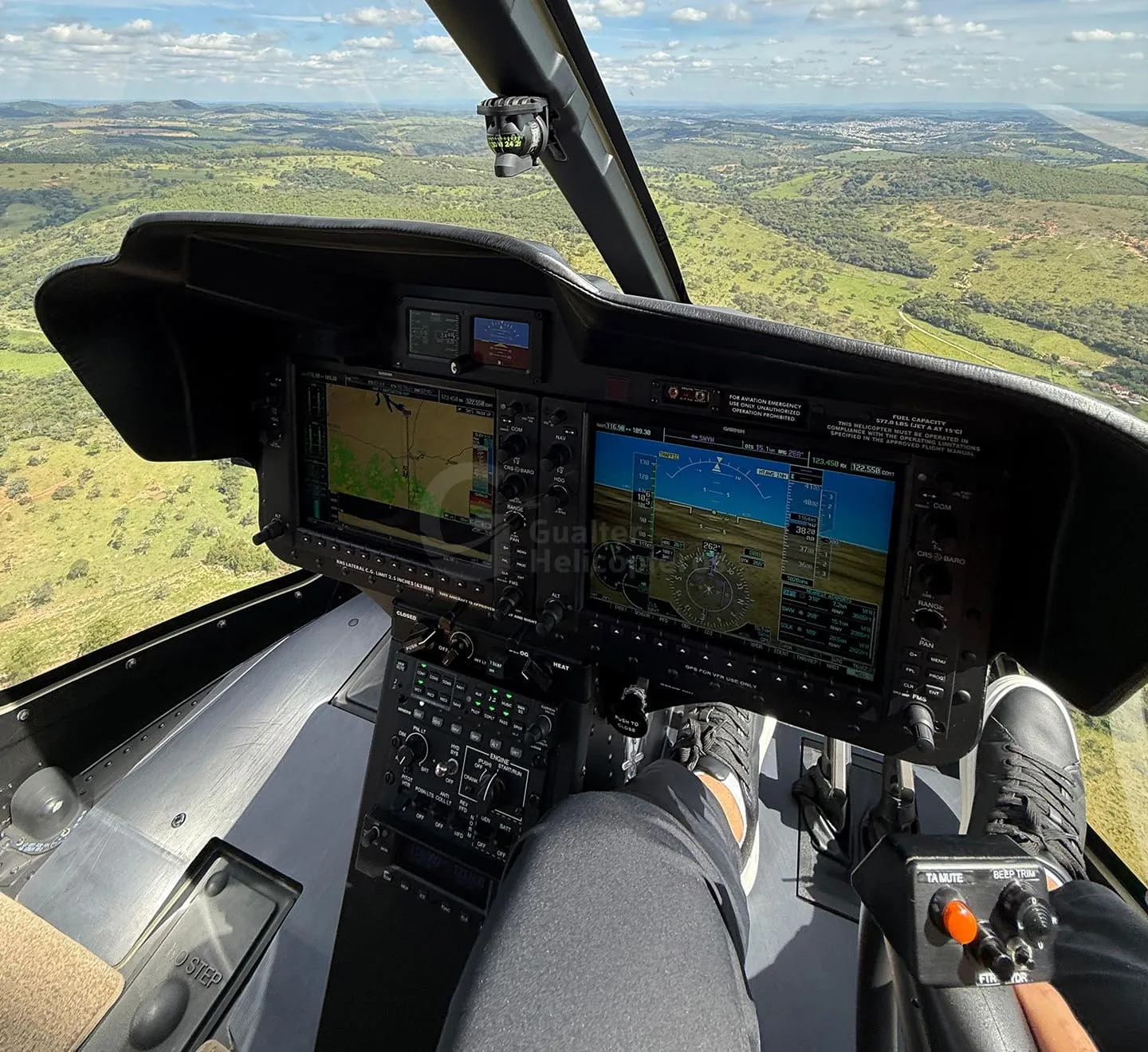 The cockpit of a helicopter shows the instrument panel with multiple digital screens and controls, with a view of green landscape outside.