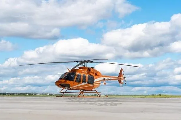 An orange Bell 505 helicopter with white stripes sits on an airfield under a partly cloudy sky.