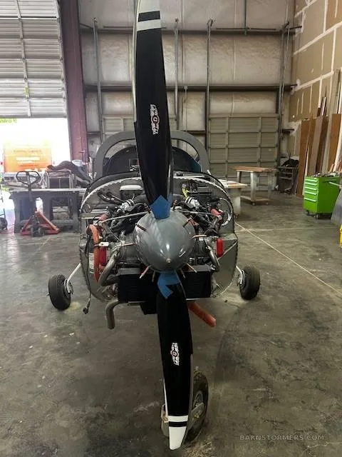 Front view of a partially assembled light aircraft with its engine exposed and a large propeller attached, in a workshop.