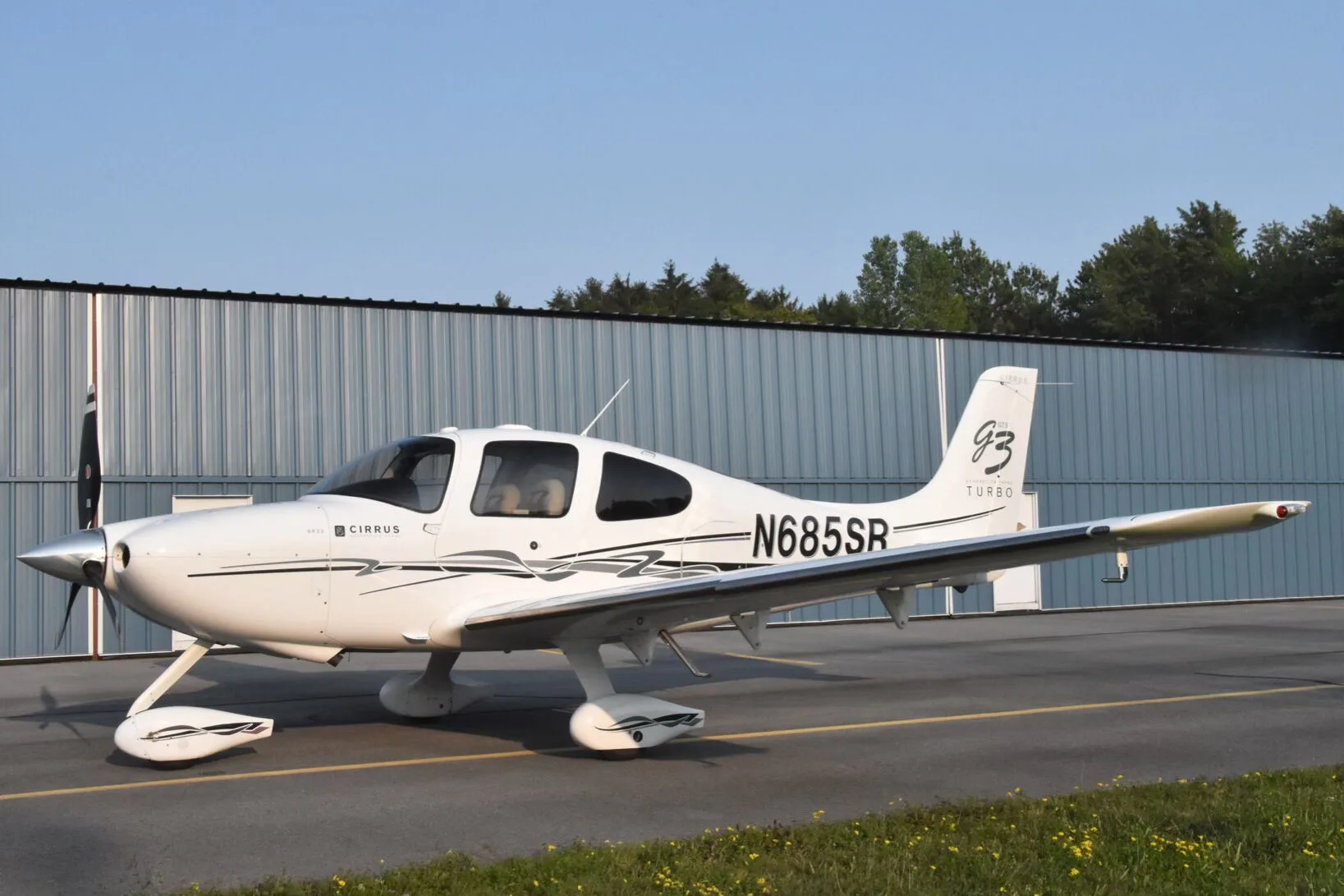 A white Cirrus SR22T aircraft (N685SR) with black and grey stripes parked on an airfield next to a hangar.