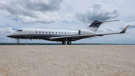 A white and dark blue business jet, likely a Bombardier Global Express, parked on a tarmac under a cloudy sky.