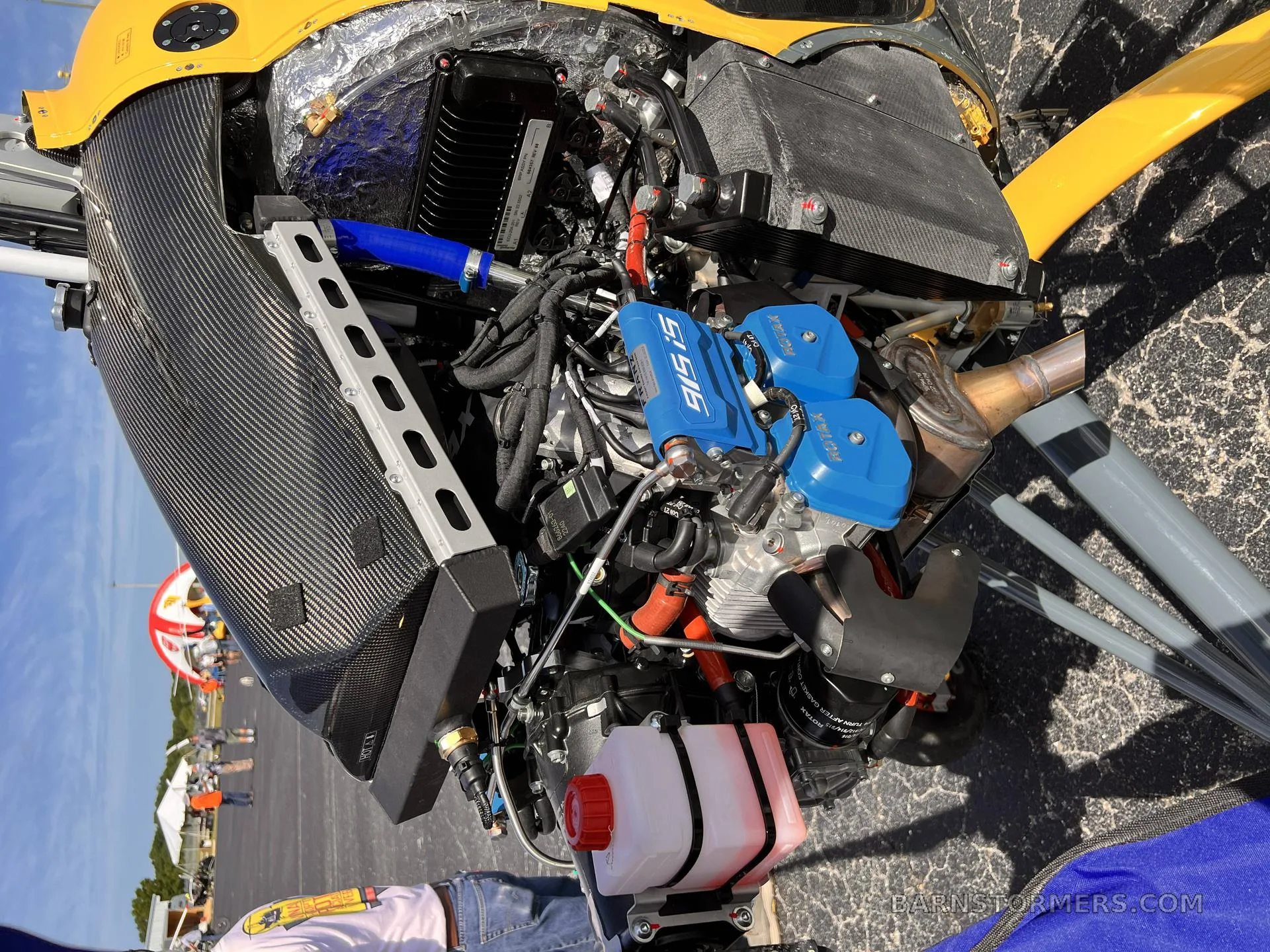 A detailed view of the engine compartment of a yellow gyrocopter, showing the blue engine covers and various mechanical components.
