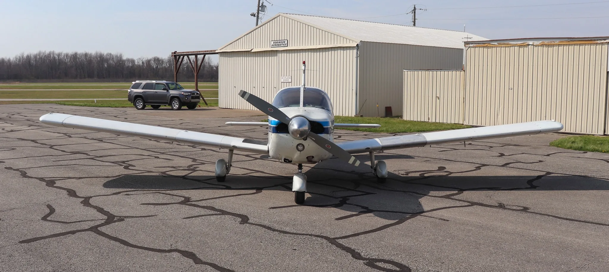A front view of the white Piper Cherokee 235 aircraft with blue stripes, parked on an asphalt ramp with hangars in the background.