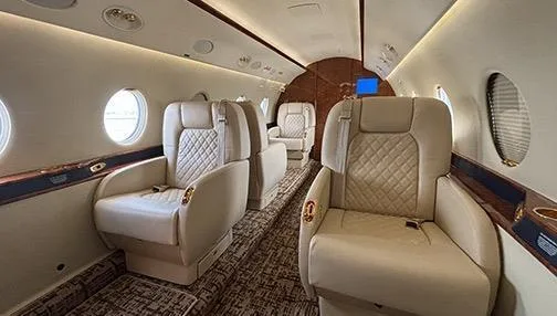 A perspective view of the private jet's cabin, showing multiple rows of beige quilted seats and oval windows.