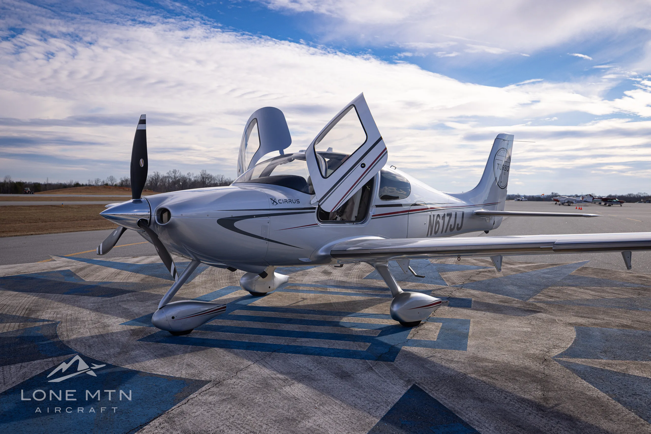 A silver Cirrus SR22T aircraft, N617JJ, with its doors open, parked on an airfield under a partly cloudy sky.