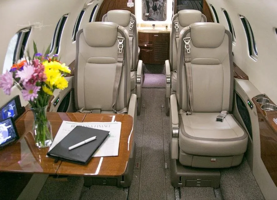 The plush interior of a private jet cabin with beige leather seats, a wooden table, and a vase of colorful flowers.
