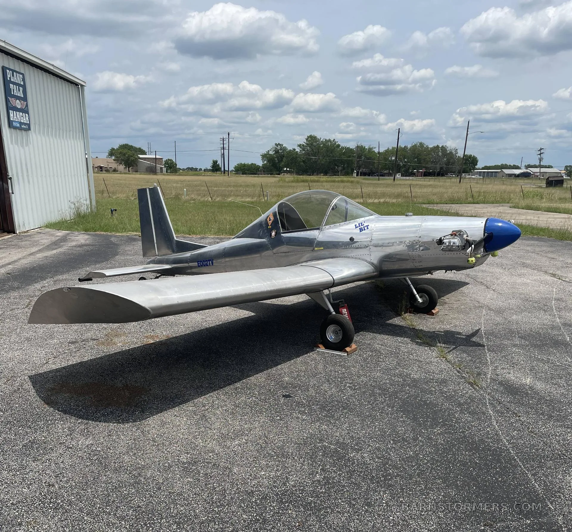 A silver single-engine aircraft with a blue nose cone is parked on an asphalt apron next to a hangar under a cloudy sky.