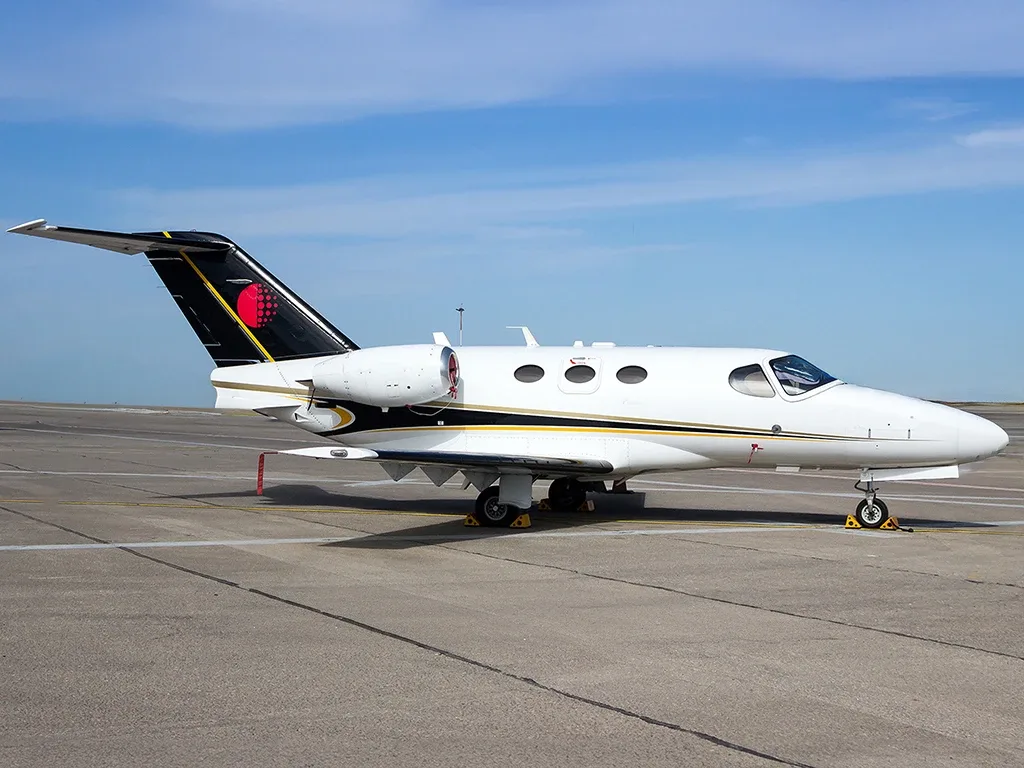 A white business jet with black and gold stripes and a red circle on the tail, parked on an asphalt tarmac under a blue sky.