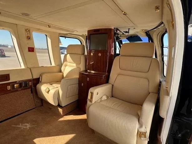 The helicopter's luxurious interior features two cream-colored captain's chairs and a wood-paneled entertainment console.