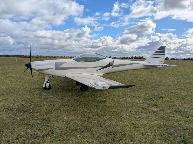 A white light aircraft with grey stripes parked on a grassy field under a cloudy sky.