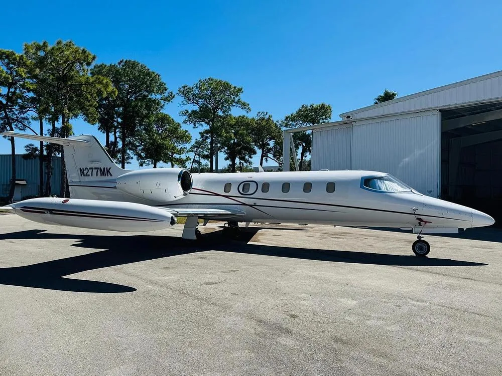 A white Learjet with a dark stripe is parked on an tarmac next to a hangar under a clear blue sky.