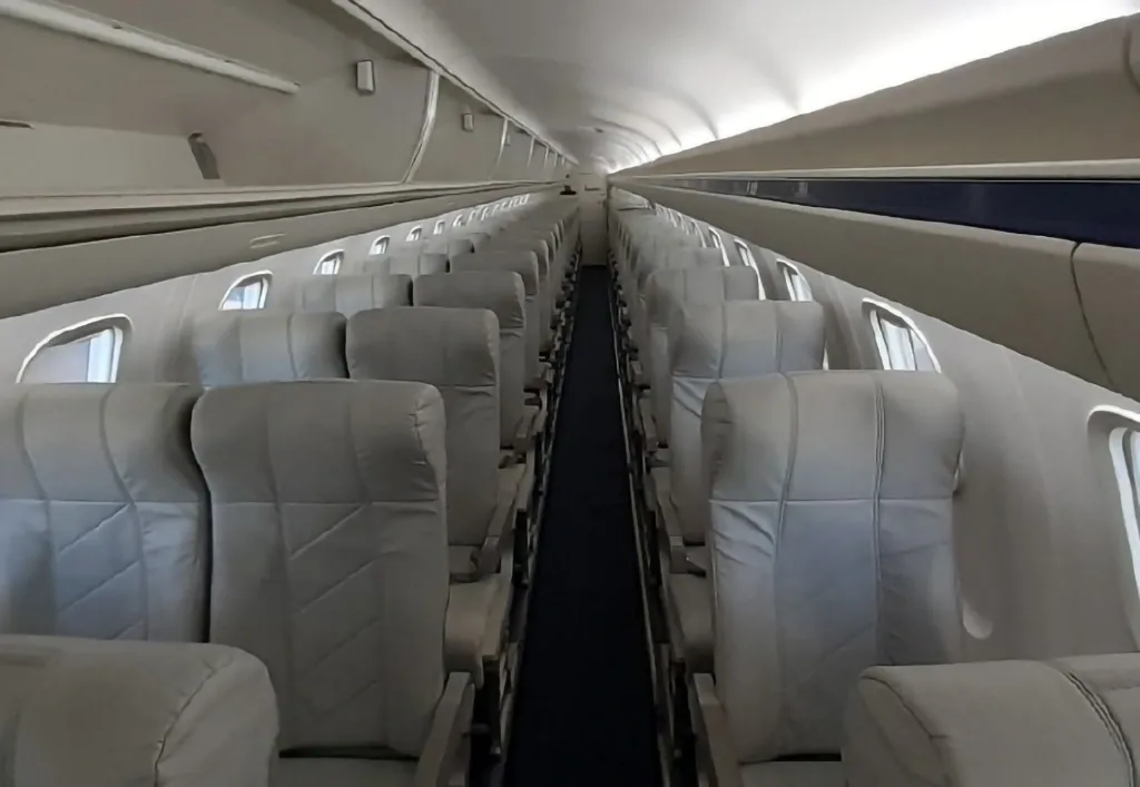 The interior of an aircraft cabin features rows of grey passenger seats along a central aisle.