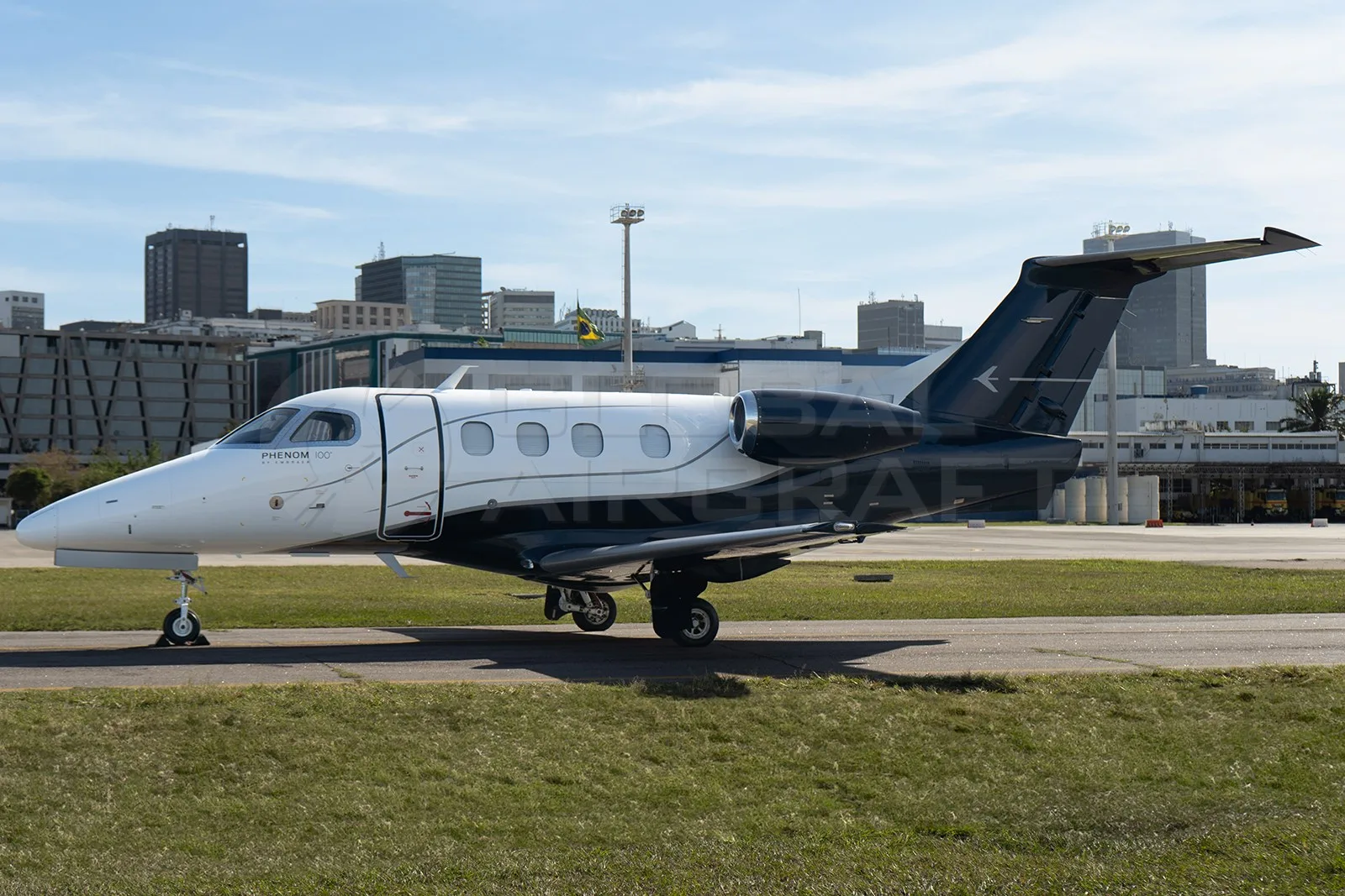 A white and black Embraer Phenom 100 business jet is parked on a tarmac with city buildings in the background.