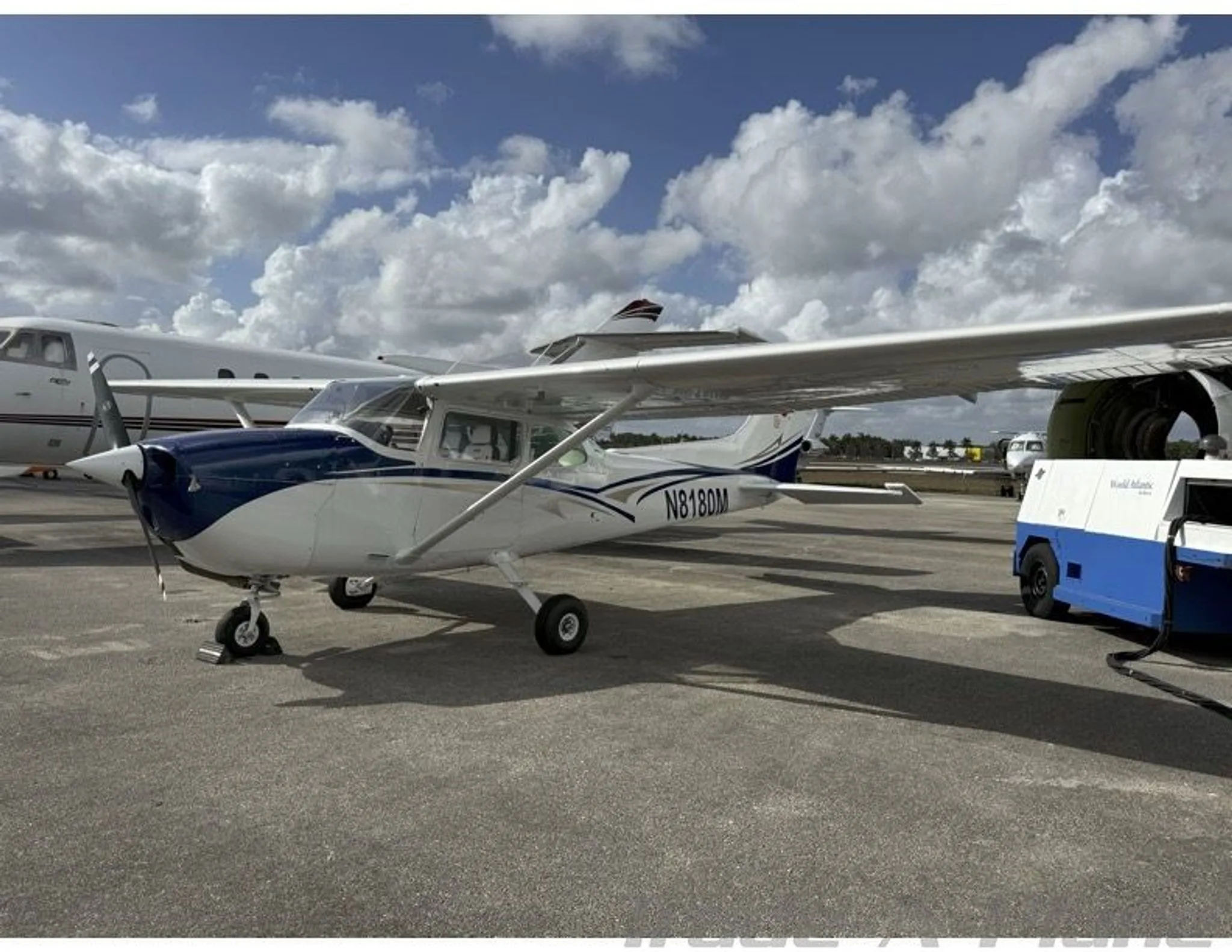 A white and blue Cessna 172 (N8180M) parked on an airfield next to a larger jet.