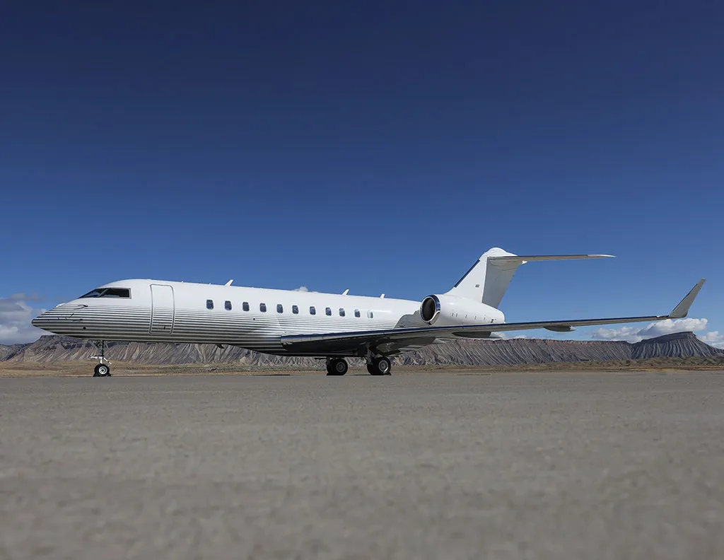 A white business jet with horizontal grey stripes on the fuselage is parked on an airfield with mountains in the background.