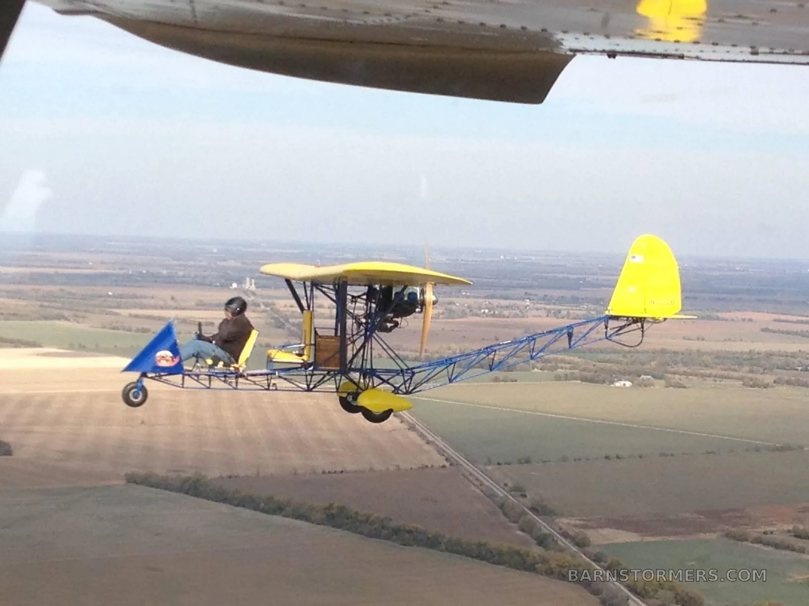 A yellow and blue ultralight aircraft with a single pilot flying over rural fields, viewed from another aircraft.