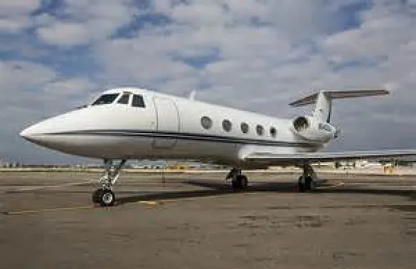 A white Gulfstream G450 business jet with blue stripes is parked on an airport tarmac under a partly cloudy sky.