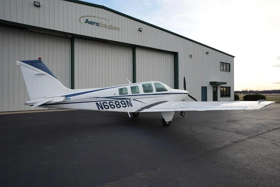 A rear-side view of the white Beechcraft Bonanza with blue and gray stripes, registration N6689N, parked on an asphalt ramp in front of a hangar.