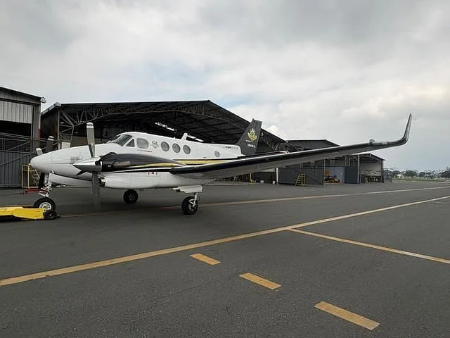 A white and black twin-engine turboprop aircraft with yellow stripes parked on an airfield next to a hangar.