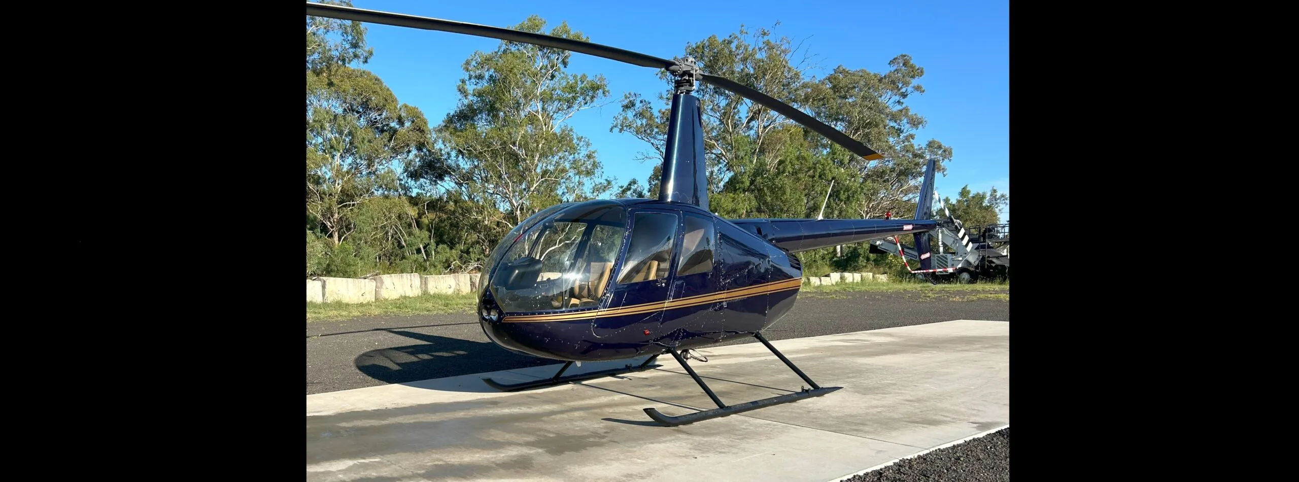 A dark blue Robinson R44 helicopter with a gold stripe on a concrete pad under a clear blue sky.