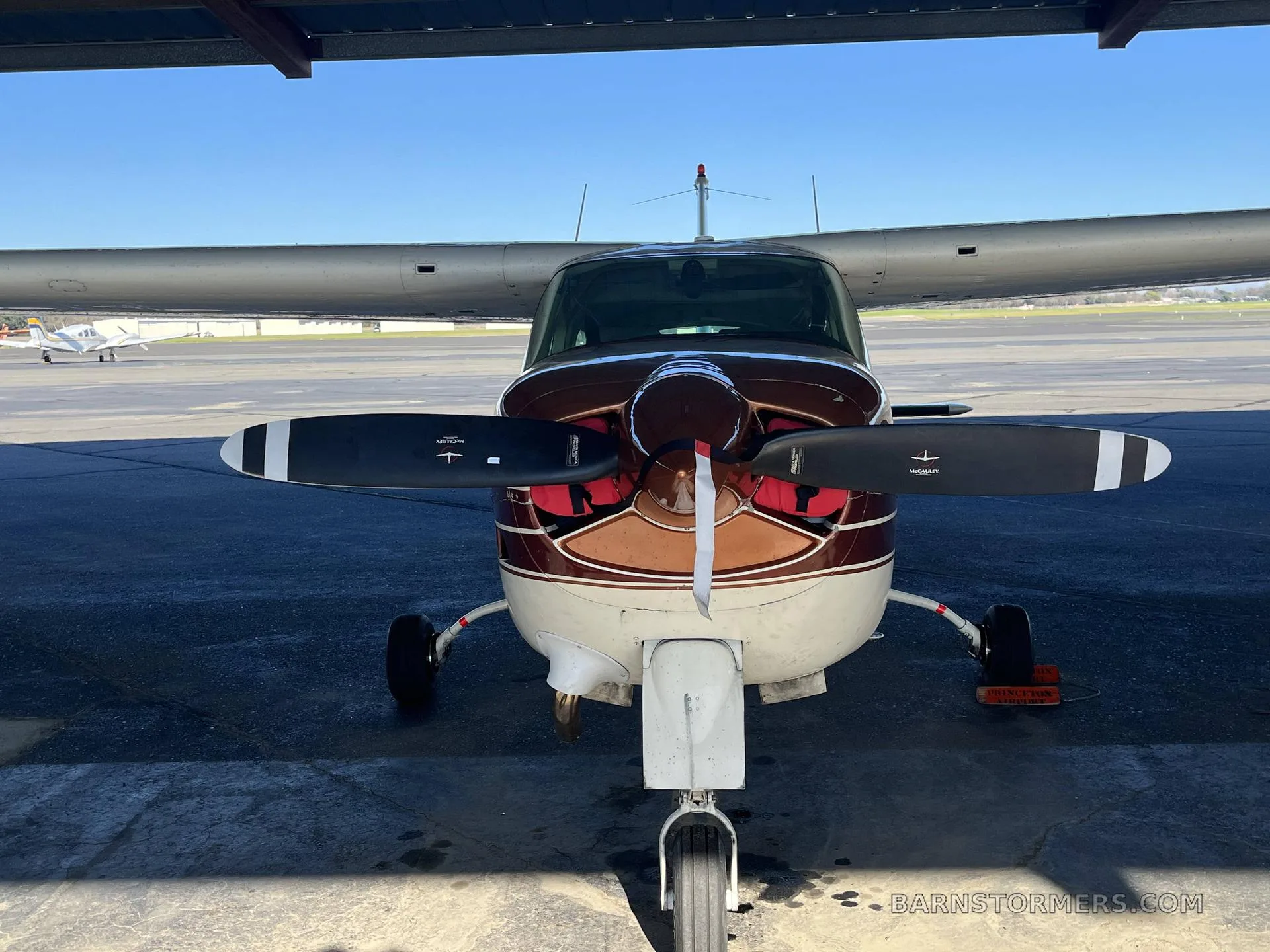 A front view of a brown and white Cessna 182 aircraft with its propeller blades visible, parked under a hangar.