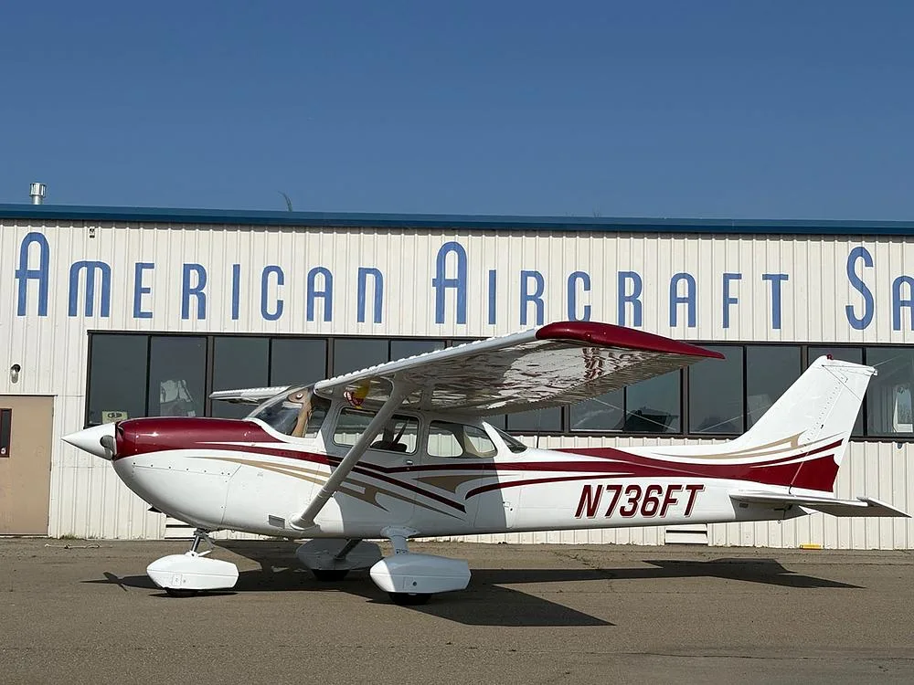 A white and maroon Cessna 172 with gold accents, parked in front of a building labeled "American Aircraft Sales".