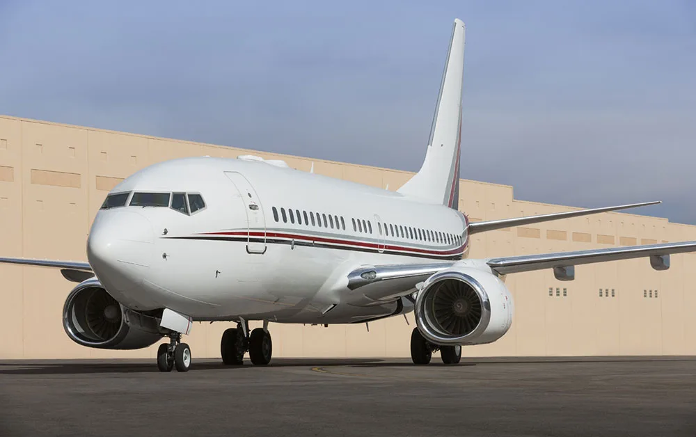 A white Boeing 737 with red and black accent stripes is parked on an airfield in front of a beige hangar.