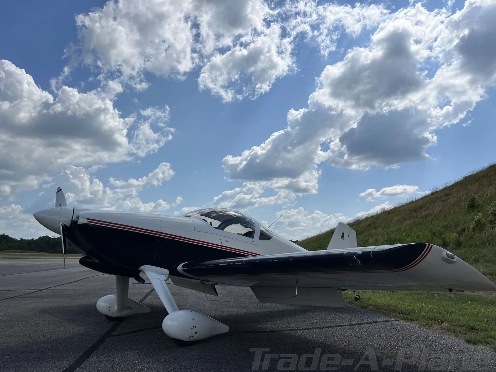 A white and dark blue single-engine aircraft with red stripes parked on an asphalt ramp under a partly cloudy sky.