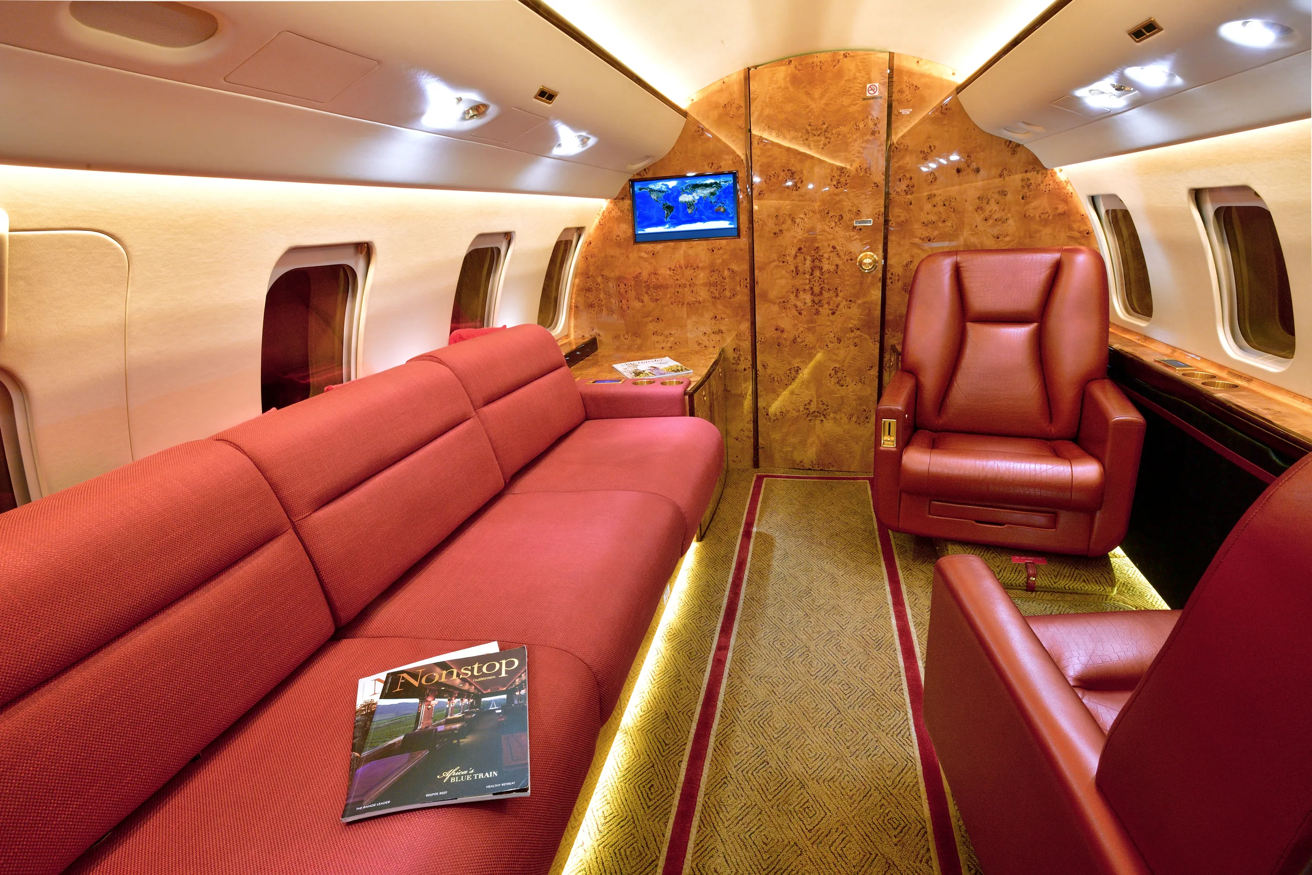 A private jet cabin interior with a long red sofa, a single red leather seat, and a wooden wall panel.
