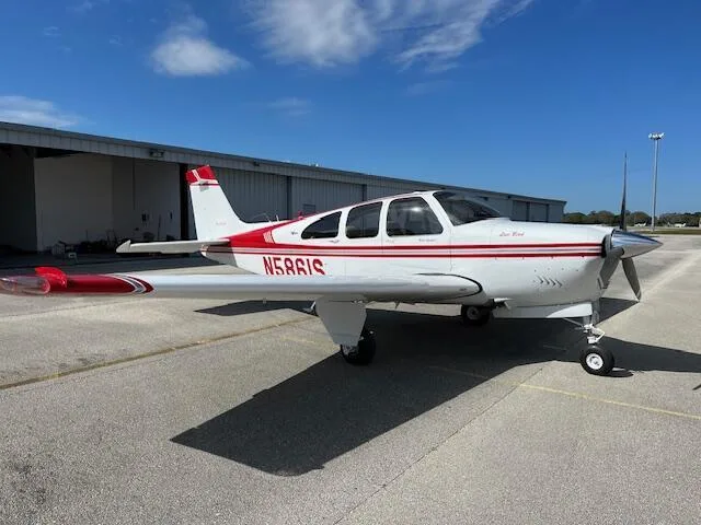 A white and red Beechcraft Bonanza N5861S parked on an asphalt ramp in front of a hangar under a blue sky.