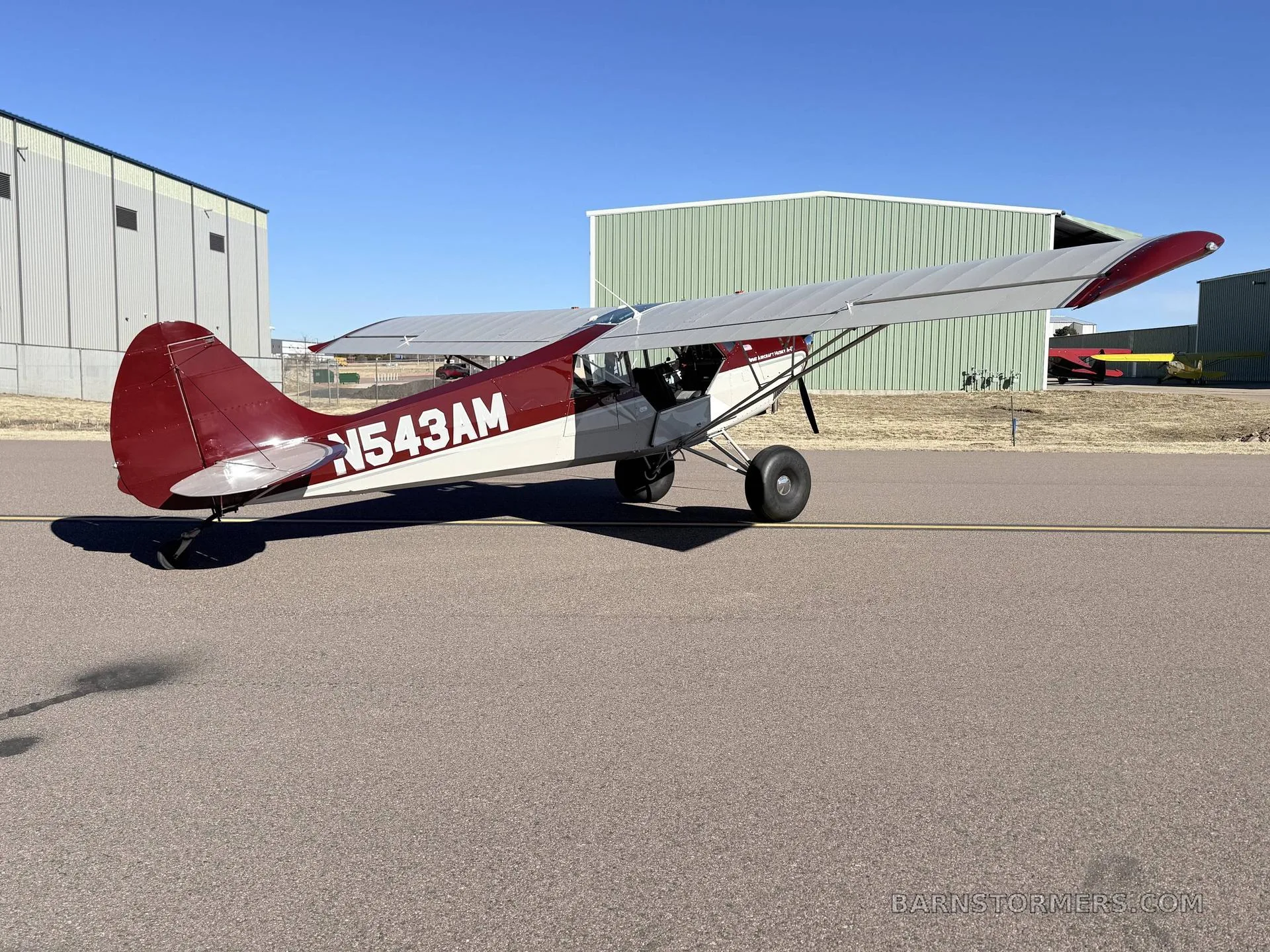A red and white Aviat Husky A-1B aircraft with large tires is seen from the rear-left on an airfield next to hangars.