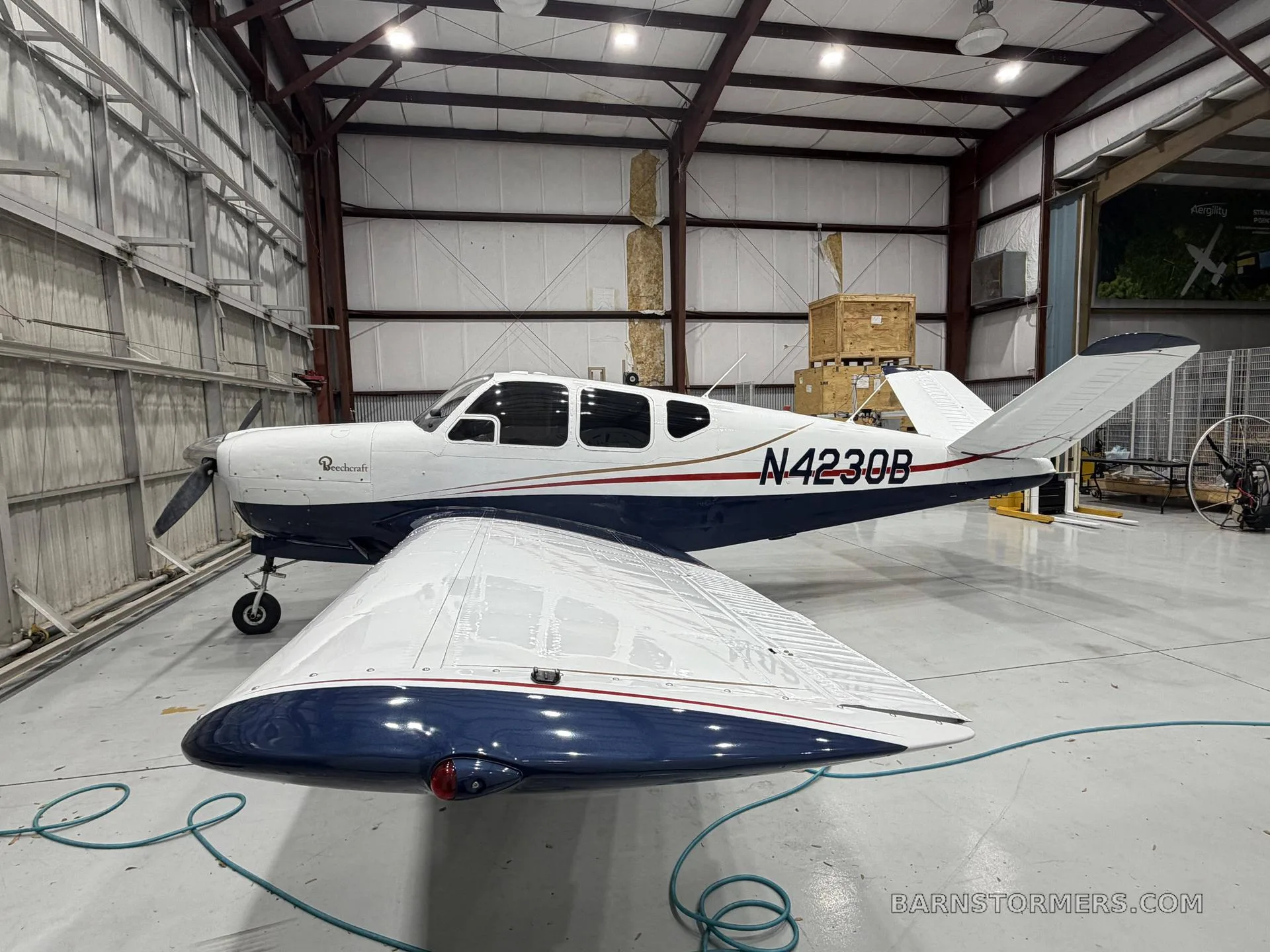 A white and blue Beechcraft Bonanza (N4230B) is parked inside a hangar, viewed from the front-right.