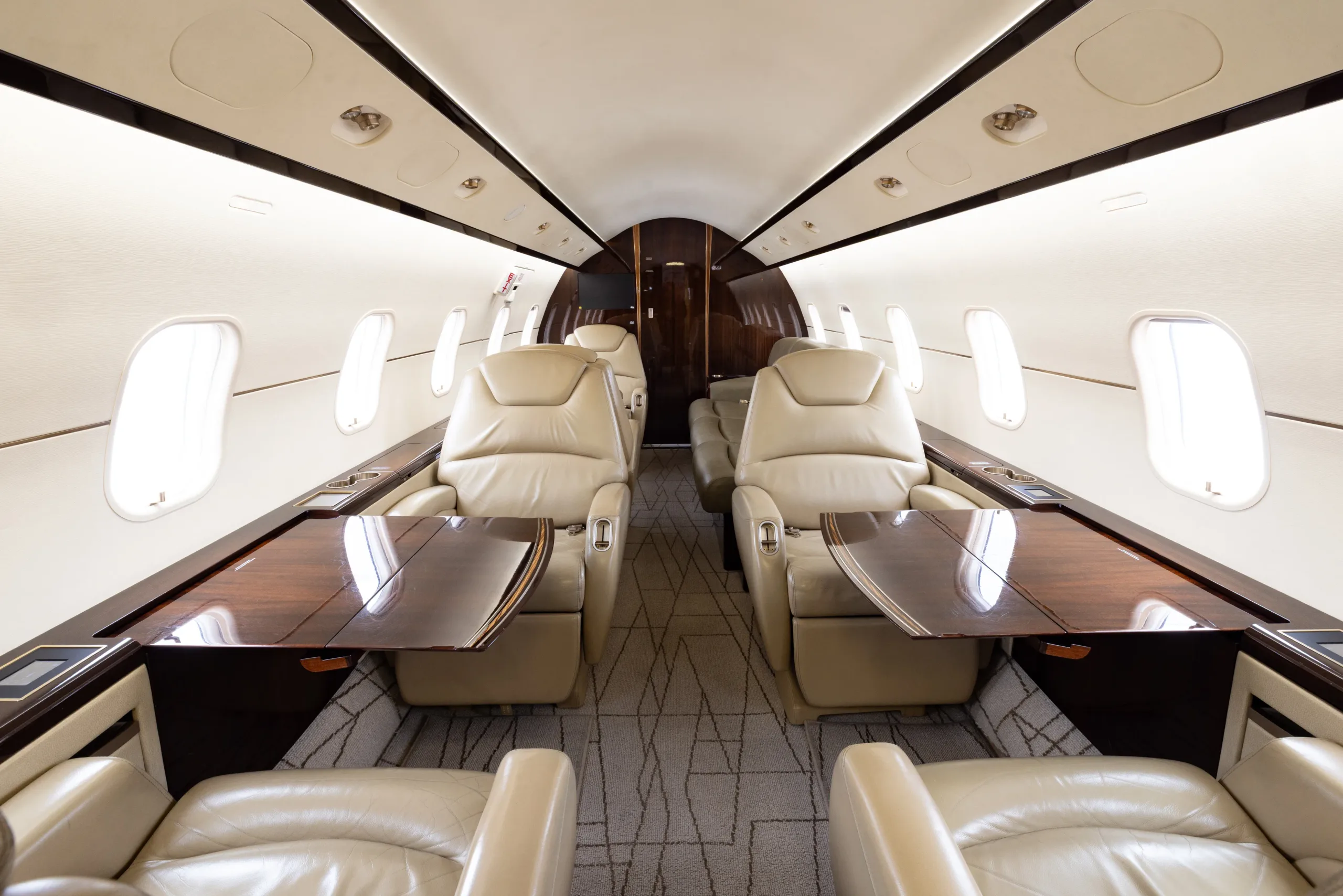 The luxurious interior of a business jet cabin with light beige leather seats and dark wood tables.