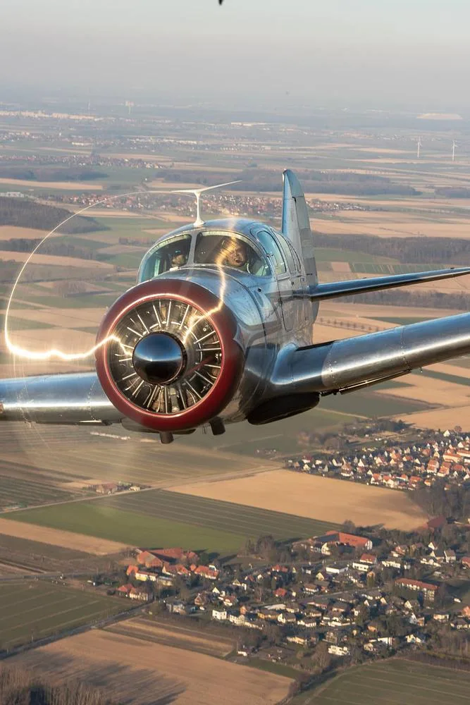 A silver vintage Beechcraft Staggerwing aircraft with a red nose cone in flight over a rural landscape.