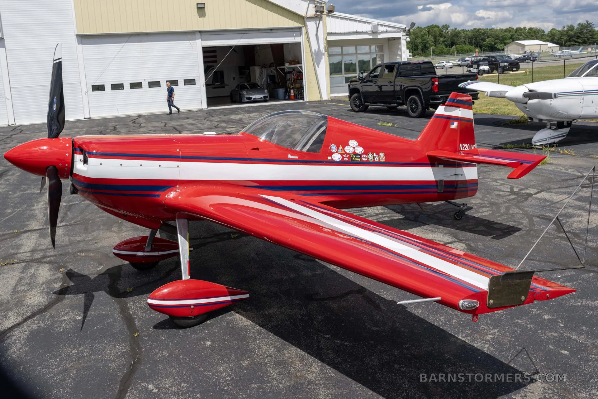 A red and white single-engine aerobatic aircraft with blue stripes, parked on an airfield outside an open hangar.
