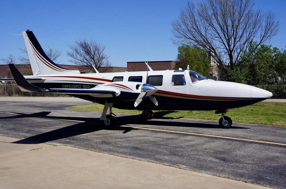 A side view of a white and dark blue twin-engine propeller aircraft with gold and red stripes, parked on an asphalt ramp.